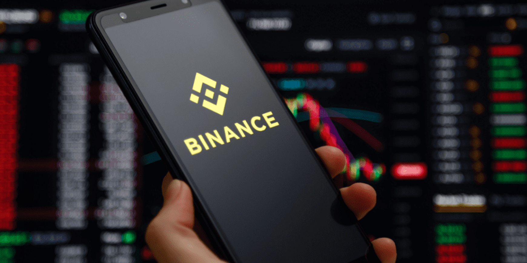 Hand houdt smartphone met het Binance-logo op het scherm, tegen een achtergrond van live cryptokoersgrafieken.