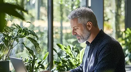 Een oudere man met grijs haar en grijs baar in een blauwe overhemd met witte rondjes die werkt op een laptop