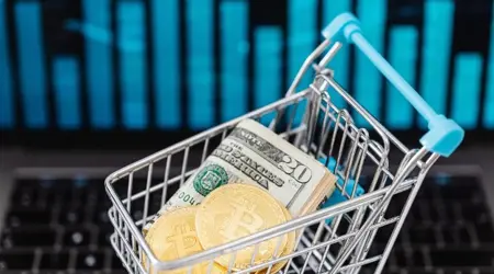 Winkelwagentje gevuld met Amerikaanse dollarbiljetten en Bitcoin-munten, op de achtergrond een laptop met grafiek.