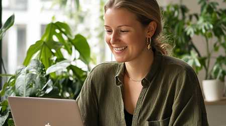 Een glimlachende vrouw die naar op een laptop aan het werk is met planten in de achtergrond.
